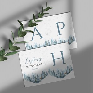 Puede incluir: Un conjunto de banderines de papel blanco con las letras azules A, P y H, y un paisaje forestal en acuarela en la parte inferior. El texto dice Easton's 1ST BIRTHDAY 20.12.202X, con copos de nieve azules e ilustraciones de ciervos.