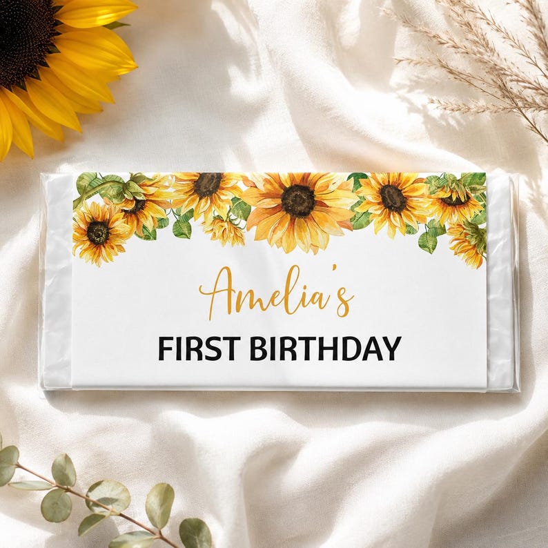 May include: A rectangular candy bar wrapper featuring a watercolor sunflower border along the top edge. The white label displays the text Amelia's in a gold script font and the words FIRST BIRTHDAY in a bold black sans-serif typeface.