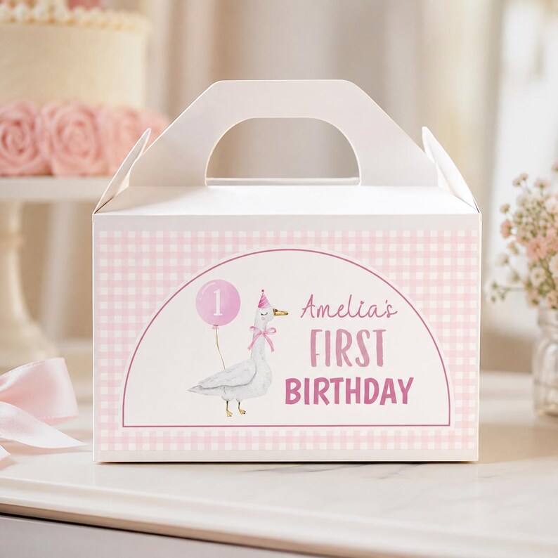 May include: A white gable box featuring a light pink gingham pattern and a central arch graphic. The design includes a watercolor illustration of a goose wearing a party hat and holding a pink balloon with the number 1, and the text Amelia's FIRST BIRTHDAY.