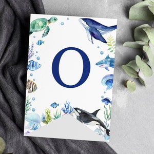 May include: A white rectangular banner featuring a large dark blue letter O in the center. The border is decorated with watercolor illustrations of various sea creatures including a sea turtle, whale, shark, orca, jellyfish, and colorful fish.