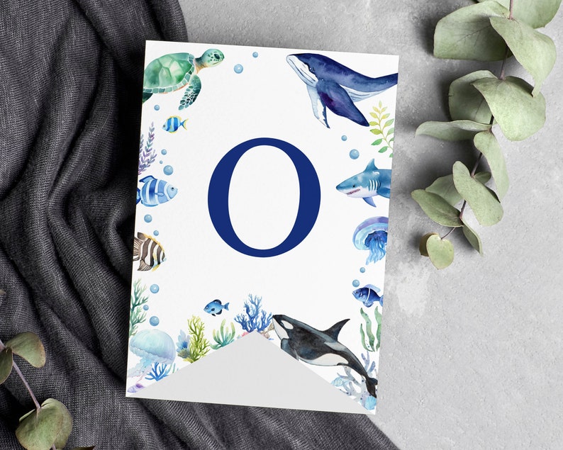 May include: A white rectangular banner featuring a large dark blue letter O in the center. The border is decorated with watercolor illustrations of various sea creatures including a sea turtle, whale, shark, orca, jellyfish, and colorful fish.