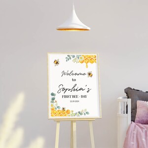 May include: A framed sign on an easel with the text "Welcome to Sophia's FIRST BEE-DAY 22.09.202X". The sign features watercolor illustrations of bees, honeycomb, and floral elements in yellow, green, and white.