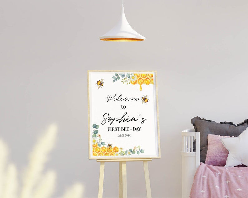 May include: A framed sign on an easel with the text "Welcome to Sophia's FIRST BEE-DAY 22.09.202X". The sign features watercolor illustrations of bees, honeycomb, and floral elements in yellow, green, and white.
