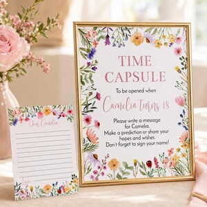May include: A gold-framed sign and a matching card featuring a colorful wildflower border. The sign reads: TIME CAPSULE To be opened when Camelia turns 18. Please write a message for Camelia. Make a prediction or share your hopes and wishes.