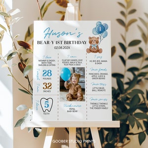 May include: A white milestone birthday sign for a first birthday featuring a brown teddy bear illustration, blue balloons, and text including Huson's Bear-y 1st Birthday, 02.08.202X, 28 pounds, 32 inches, 5 teeth, and a photo of a child in a bear costume.