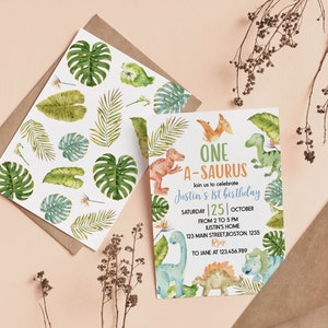 Op de afbeelding: Een verjaardagsuitnodigingskaart en een bijpassende envelopvoering met aquarelillustraties van diverse groene tropische bladeren en kleurrijke dinosaurussen. De tekst luidt ONE A-SAURUS, join us to celebrate Justin's 1st birthday, Saturday 25 October.