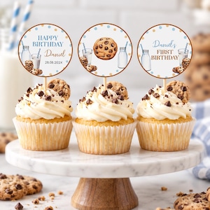 May include: Three cupcakes with white frosting and chocolate chips, each topped with a circular paper topper. The toppers feature illustrations of milk bottles, cookies, and text including Happy Birthday, Daniel, and First Birthday with the date 28.08.202X.