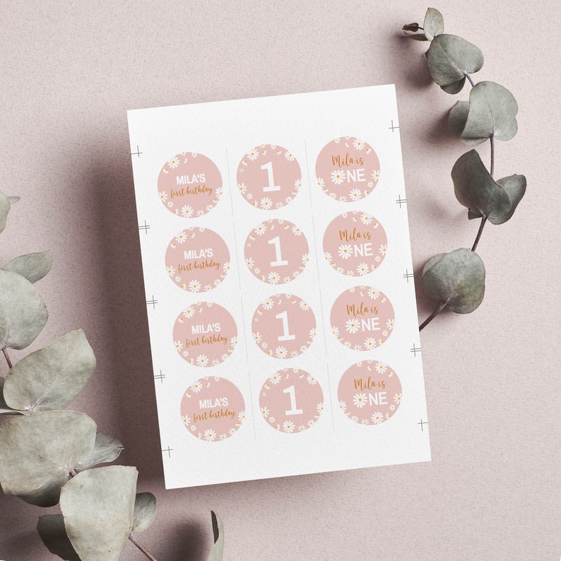 May include: A sheet of twelve circular stickers with a dusty pink background and white daisy pattern. The stickers feature the text Mila's first birthday, the number 1, and the text Mila is ONE in white and gold script and block lettering.