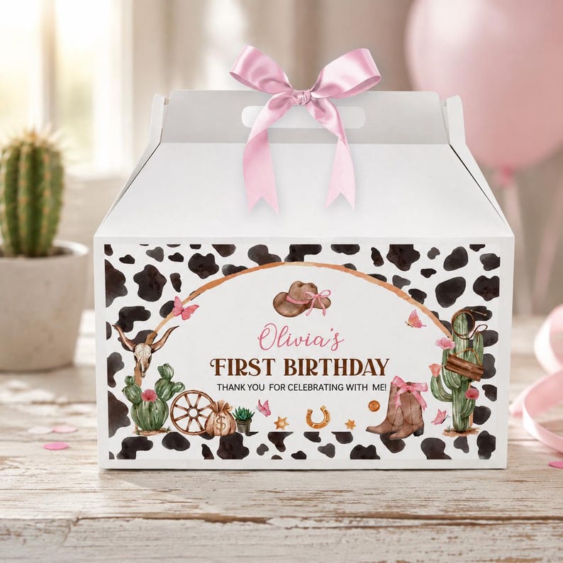 May include: A white gable box with a pink satin ribbon bow on top, featuring a black and white cow print pattern. The front displays a western-themed graphic with a cowboy hat, boots, cacti, a wagon wheel, and the text Olivia's FIRST BIRTHDAY.