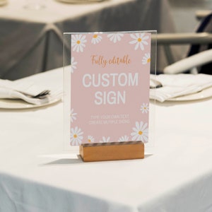 May include: A rectangular acrylic sign in a wooden base featuring a light pink background with white daisy illustrations. The text reads: Fully editable, CUSTOM SIGN, TYPE YOUR OWN TEXT, CREATE MUTIPLE SIGNS.