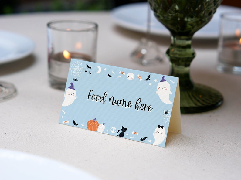 May include: A light blue folded card with the text Food name here in black script. The card features a Halloween-themed border with illustrations of white ghosts wearing purple hats, orange pumpkins, a black cat, spider webs, bats, and candy.