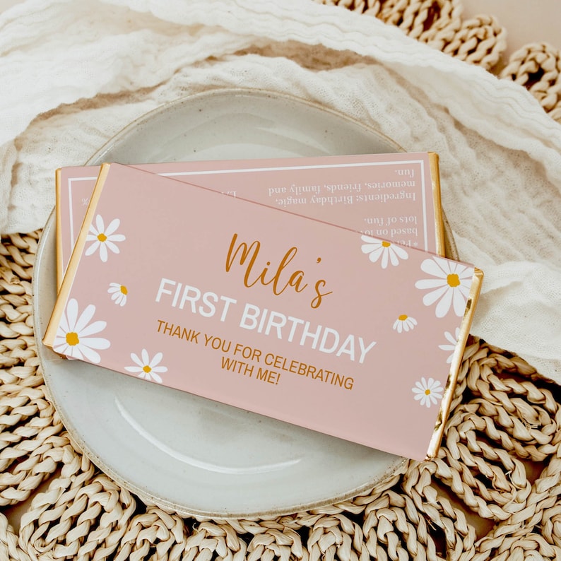 Op de afbeelding: Roze en gouden chocoladeverpakking met witte madeliefjes. De verpakking zegt "Mila's First Birthday" en "Bedankt voor het vieren met me!"