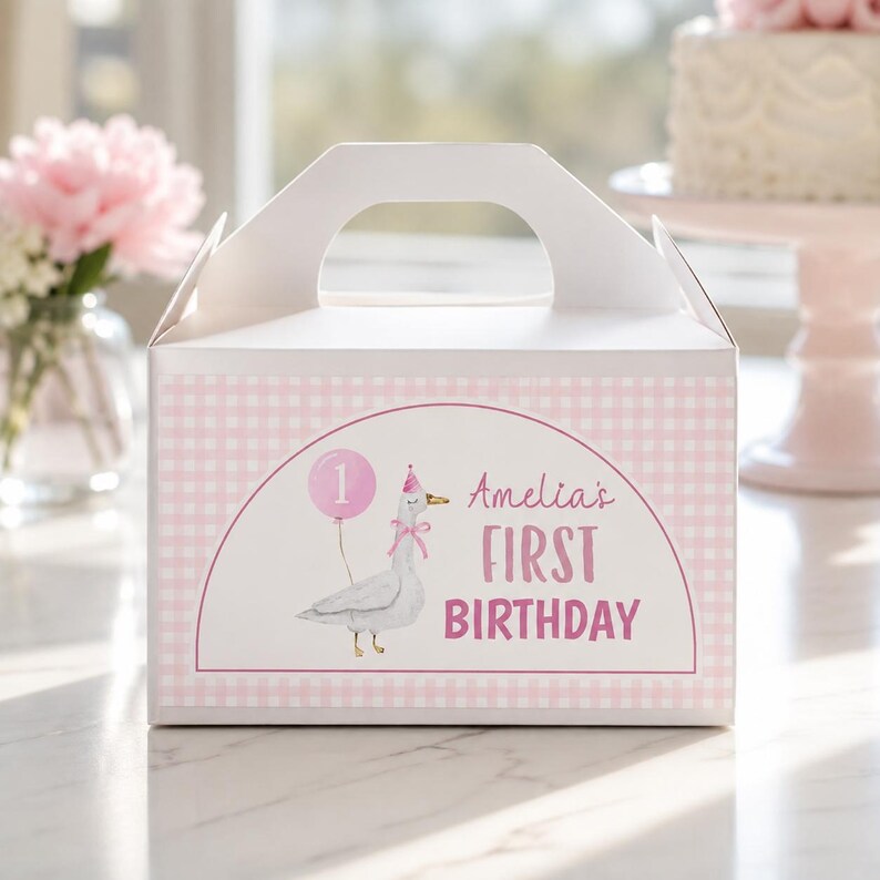 May include: A white cardboard gable box featuring a light pink gingham pattern. The front displays a graphic of a goose wearing a party hat and holding a pink balloon with the number 1, alongside the text Amelia's FIRST BIRTHDAY in pink.