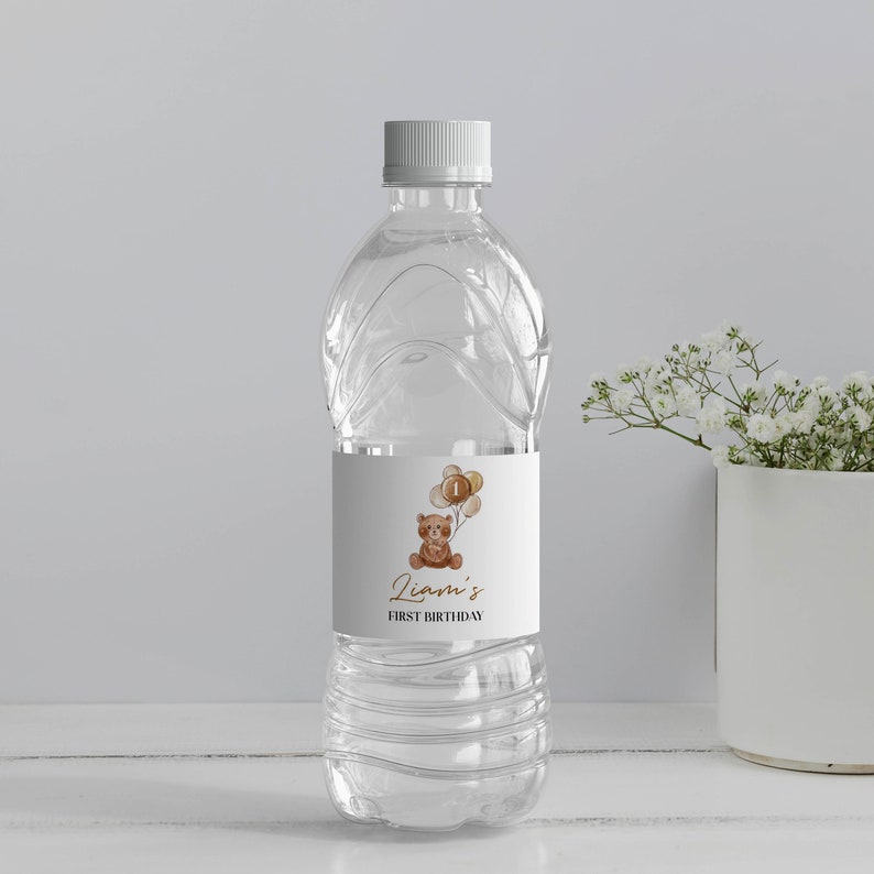 May include: A clear plastic water bottle with a white label featuring a watercolor illustration of a brown teddy bear holding a cluster of tan balloons with the number one on the center balloon. The text reads Liam's FIRST BIRTHDAY.