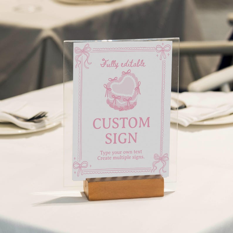 Peut inclure: Un porte-affiche en acrylique transparent avec une base en bois pr&eacute;sentant une carte blanche avec du texte et des graphismes roses. La carte comporte une illustration de g&acirc;teau en forme de c&oelig;ur, des n&oelig;uds roses et le texte : Fully editable, CUSTOM SIGN, Type your own text, Create multiple signs.