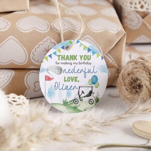 May include: A round white gift tag with a hole at the top and a jute string. The tag features a watercolor golf theme with a golf cart, a flag, and balloons. Text reads: THANK YOU for making my birthday onederful LOVE, Oliver in blue and green.