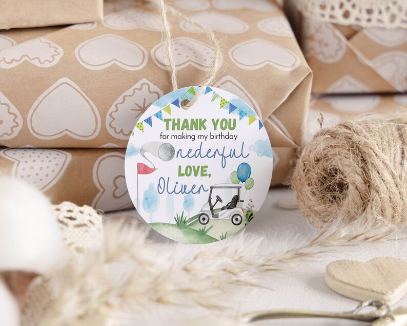 May include: A round white gift tag with a hole at the top and a jute string. The tag features a watercolor golf theme with a golf cart, a flag, and balloons. Text reads: THANK YOU for making my birthday onederful LOVE, Oliver in blue and green.