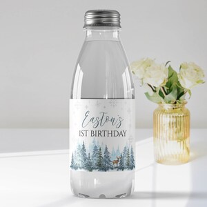 May include: A clear plastic water bottle with a silver screw cap. The label features a winter forest scene with blue pine trees, a small deer, and snowflakes. The text reads Easton's 1ST BIRTHDAY in blue and black lettering.
