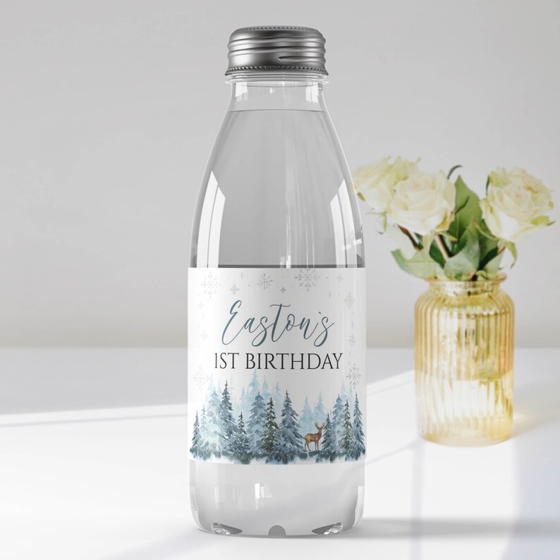 May include: A clear plastic water bottle with a silver screw cap. The label features a winter forest scene with blue pine trees, a small deer, and snowflakes. The text reads Easton's 1ST BIRTHDAY in blue and black lettering.