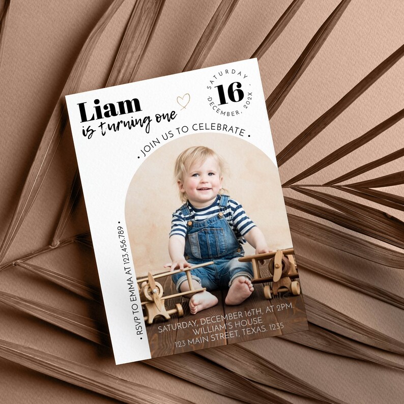 May include: A rectangular birthday invitation card featuring a photo of a child in denim overalls and a striped shirt with two wooden toy airplanes. Text includes Liam is turning one, Saturday 16 December 202X, Join us to celebrate, and RSVP details.