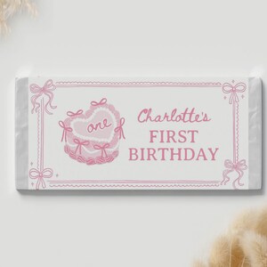 May include: A rectangular candy bar wrapper featuring a white background with pink line art. The design includes a heart-shaped cake with the word one, and the text Charlotte's FIRST BIRTHDAY, framed by decorative pink bows and borders.