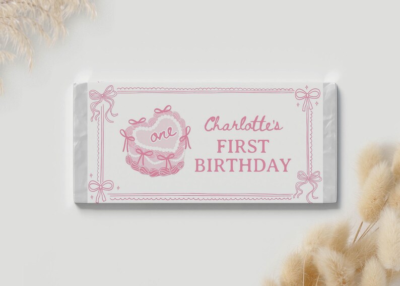 May include: A rectangular candy bar wrapper featuring a white background with pink line art. The design includes a heart-shaped cake with the word one, and the text Charlotte's FIRST BIRTHDAY, framed by decorative pink bows and borders.