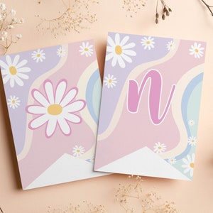 Op de afbeelding: Twee rechthoekige wimpels met een golvende pastelachtergrond van lavendel, roze en lichtblauw. De ene vlag toont een grote witte margriet met een geel hart, terwijl de andere een grote roze kleine letter n bevat.
