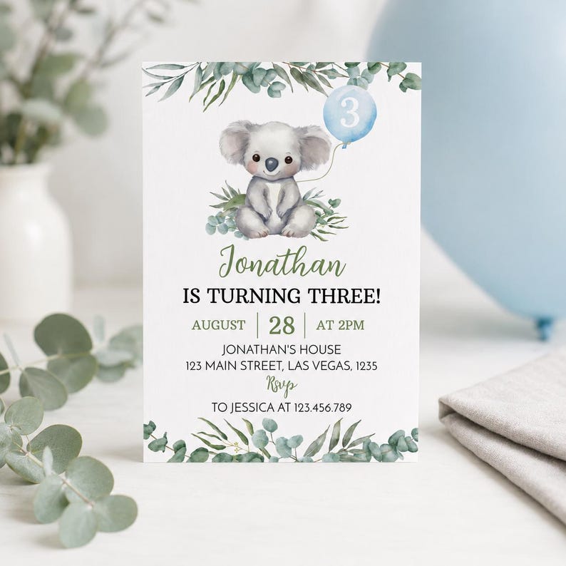 May include: A rectangular birthday invitation card featuring a watercolor illustration of a koala holding a light blue balloon with the number 3. The card includes green eucalyptus foliage at the top and bottom, with text detailing Jonathan's third birthday party information.