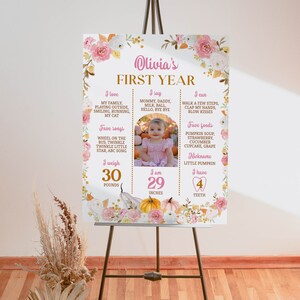 May include: A rectangular milestone sign titled Olivia's FIRST YEAR featuring pink and white floral borders, pumpkin illustrations, and a central photo of a child. It lists personal details including 30 pounds, 29 inches, and 4 teeth in gold and pink text.