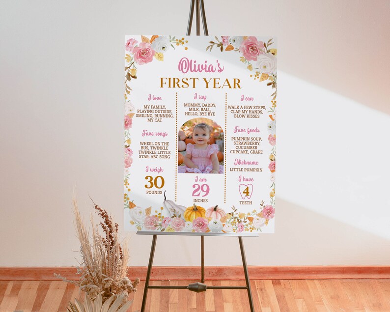 May include: A rectangular milestone sign titled Olivia's FIRST YEAR featuring pink and white floral borders, pumpkin illustrations, and a central photo of a child. It lists personal details including 30 pounds, 29 inches, and 4 teeth in gold and pink text.