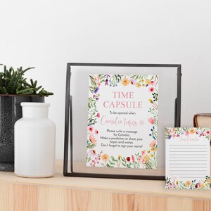 May include: A rectangular sign in a black frame and a matching small lined card, both featuring a colorful watercolor floral border. The sign reads: TIME CAPSULE To be opened when Camelia turns 18. Please write a message for Camelia.