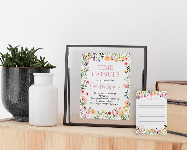 May include: A rectangular sign in a black frame and a matching small lined card, both featuring a colorful watercolor floral border. The sign reads: TIME CAPSULE To be opened when Camelia turns 18. Please write a message for Camelia.