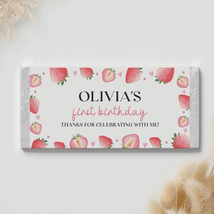 May include: A rectangular candy bar wrapper featuring a white background with a border of watercolor strawberries and small pink hearts. The text reads OLIVIA'S first birthday and THANKS FOR CELEBRATING WITH ME in black and pink lettering.