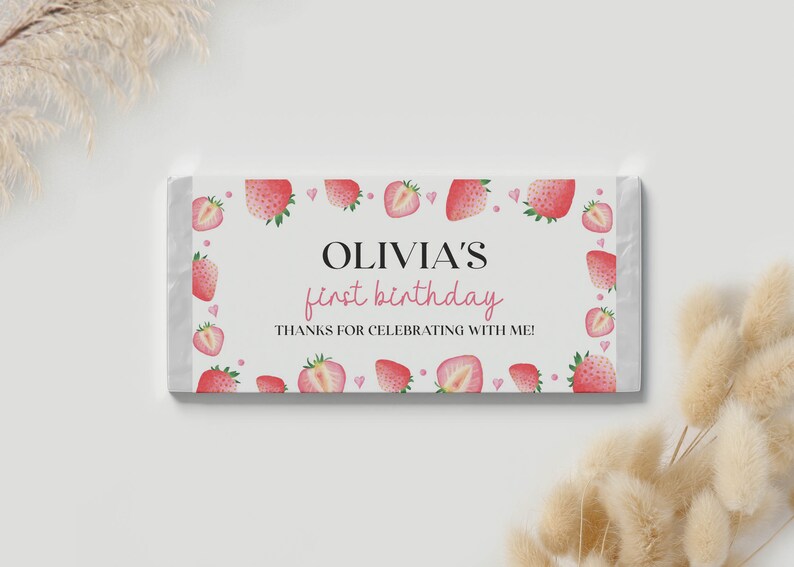 May include: A rectangular candy bar wrapper featuring a white background with a border of watercolor strawberries and small pink hearts. The text reads OLIVIA'S first birthday and THANKS FOR CELEBRATING WITH ME in black and pink lettering.
