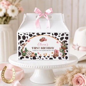 May include: A white gable box with a pink satin ribbon bow on top, featuring a black and white cow print pattern. The front displays a watercolor graphic with a cowboy hat, cactus, horseshoe, and the text Olivia's FIRST BIRTHDAY THANK YOU.