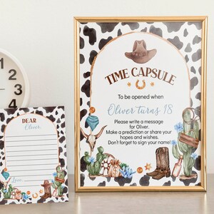 May include: A printable time capsule sign with a cowboy theme. The sign has a brown and white cow print background with a gold frame. The text on the sign reads "Time Capsule" with a horseshoe symbol and "To be opened when Oliver turns 18". There are illustrations of a cowboy hat, boots, cacti, and a skull.