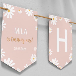 Op de afbeelding: Een decoratieve slinger met lichtroze vlaggetjes voorzien van witte madeliefjespatronen. E&eacute;n vlaggetje toont de tekst MILA is turning one! 10.08.202X in witte en gouden letters. Andere vlaggetjes tonen de grote witte letters H en A, geregen aan een bruin koord.