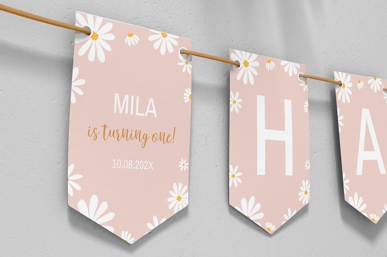 Op de afbeelding: Een decoratieve slinger met lichtroze vlaggetjes voorzien van witte madeliefjespatronen. E&eacute;n vlaggetje toont de tekst MILA is turning one! 10.08.202X in witte en gouden letters. Andere vlaggetjes tonen de grote witte letters H en A, geregen aan een bruin koord.