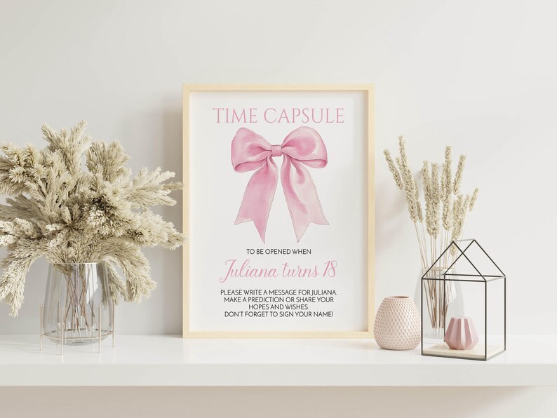 May include: A white frame with a pink bow and the text "TIME CAPSULE" at the top. The text below the bow reads "TO BE OPENED WHEN Juliana turns 18".  The text below that reads "PLEASE WRITE A MESSAGE FOR JULIANA MAKE A PREDICTION OR SHARE YOUR HOPES AND WISHES DON'T FORGET TO SIGN YOUR NAME".