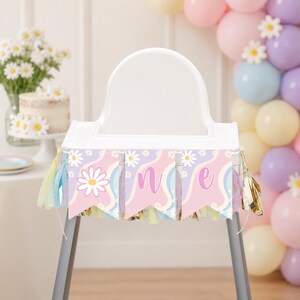 Op de afbeelding: Een kinderstoelbanner met drie wimpels op een golvende roze, paarse en witte achtergrond, versierd met witte madeliefjes. De banner toont het woord ONE in een cursief lettertype, geaccentueerd met gouden en lichtblauwe franjes.