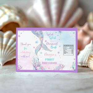 May include: A rectangular card with a purple border featuring a watercolor mermaid tail, starfish, and coral illustrations. Text reads: Splish Splash It's a Mermaid Bash, Mayra's FIRST BIRTHDAY, and Thank you for celebrating with me, plus a nutrition label.