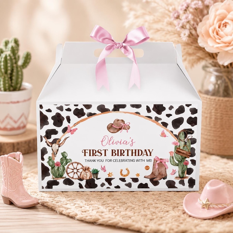 May include: A white cardboard gable box with a pink satin ribbon bow on top. The box features a black and white cow print pattern and a central graphic with a brown cowboy hat, cacti, a skull, a wagon wheel, and the text Olivia's FIRST BIRTHDAY.