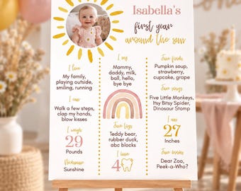 May include: A white milestone birthday sign titled Isabella's first year around the sun, featuring a photo of a baby, a yellow sun graphic, a watercolor rainbow, and text detailing favorite foods, songs, books, toys, weight of 29 pounds, and height of 27 inches.