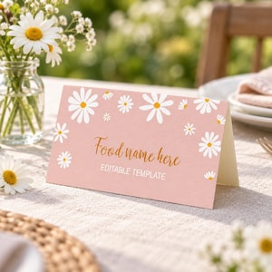 Op de afbeelding: Een gevouwen roze kaart met een wit madeliefjespatroon met de gouden cursieve tekst Food name here en de hoofdlettertekst EDITABLE TEMPLATE. De kaart staat op een gestructureerd oppervlak met een glazen vaas met witte madeliefjes.