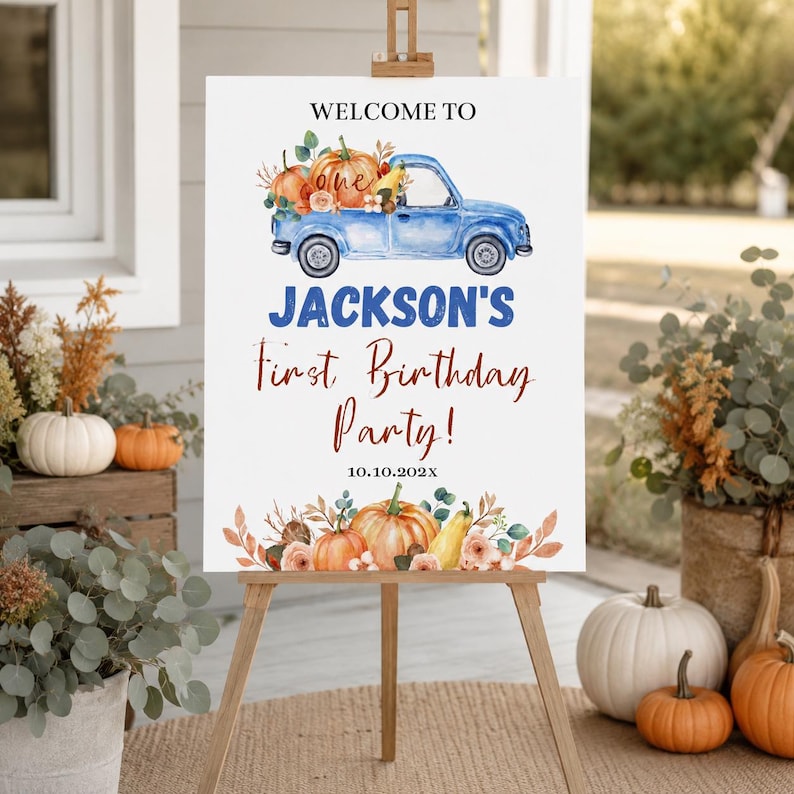May include: A white rectangular sign on a wooden easel featuring a blue truck illustration, orange pumpkins, and text reading WELCOME TO JACKSON'S First Birthday Party! 10.10.202X. The design includes watercolour-style floral and pumpkin accents.
