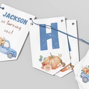 May include: A decorative party banner featuring white pennants strung on a blue cord. The pennants display the text JACKSON is turning one! in blue and brown, alongside watercolor illustrations of a blue truck carrying orange pumpkins.