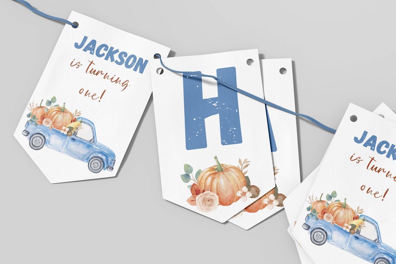May include: A decorative party banner featuring white pennants strung on a blue cord. The pennants display the text JACKSON is turning one! in blue and brown, alongside watercolor illustrations of a blue truck carrying orange pumpkins.