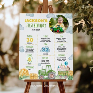 May include: A colorful first birthday poster for a boy named Jackson. The poster features a photo of the boy in a green tractor, along with details about his age, weight, height, favorite foods, toys, and songs. The poster is decorated with a green tractor, farm animals, and a blue sky with white clouds.
