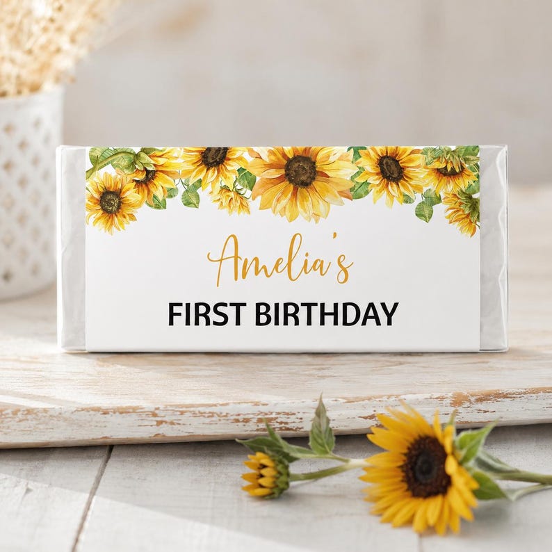 May include: A rectangular candy bar wrapper featuring a white background with a border of yellow sunflowers and green leaves along the top edge. The text "Amelia's" is printed in a gold script font above the black text "FIRST BIRTHDAY".