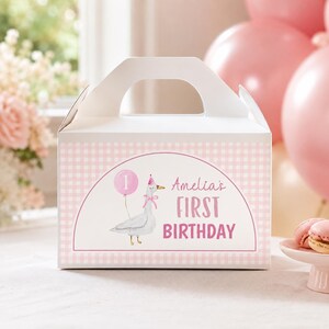 May include: A white gable box featuring a light pink gingham pattern. The front displays a semi-circular label with a watercolor illustration of a goose wearing a pink party hat and holding a pink balloon with the number 1, and the text Amelia's FIRST BIRTHDAY.